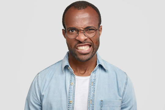 Irritated African American guy frowns face in displeasure, being annoyed with bad news, clenches teeth, wears fashionable denim shirt and glasses, isolated over white background. Negative emotions