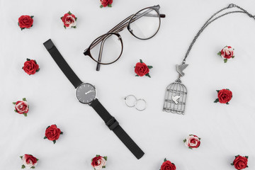 Eyeglasses, black watches, necklace and rings decorate with red rose paper flowers on white fabric 