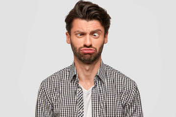 Funny European male makes grimace, purses lips and crosses eyes, foolishes indoor, has fun, dressed in fashionable shirt, isolated over white background. People and facial expressions concept