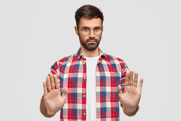 Pleasant looking serious male office worker refuses to help with preparing project, shows stop gesture, asks not bother him during vacation, has thick beard and mustache, isolated on white wall