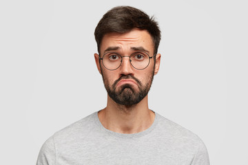 Headshot of attractive young male with beard, purses lips, feels puzzled, has hesitant and miserable look, being disappointed and displeased, dressed casually, isolated over white background. - obrazy, fototapety, plakaty