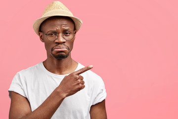 Displeased African American male has sorrowful dissatisfied expression, shows with fore finger...