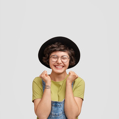 Joyful European female in black hat and denim dungarees, clenches fists and has broad smie, being in high spirit after successful day, poses against white background. People and happiness concept