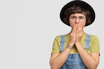 Studio shot of angry female with surprised dissatisfied expression, covers mouth with palms, being shocked by failure, doesn`t expect to recieve bad comments about her work, stands against white wall