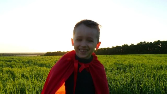 A happy child in a superhero costume in a red raincoat runs along a green lawn against the backdrop of a sunset in slow motion
