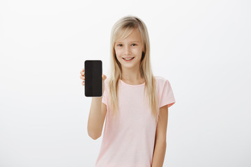 Studio shot of intrigued neat blond girl in pink t-shirt, smiling curiously and showing black smartphone at camera, asking brother password from device, wanting play games or brows in network