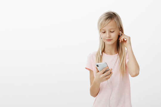 Smart girl knows everything about gadgets. Portrait of cute beautiful blond young child, wearing earphones and picking song in smartphone, looking at screen focused, standing over gray background