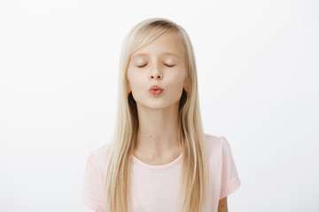 Girl practising near mirror how to kiss. Portrait of cute fashionable young girl with blond hair, folding lips and closing eyes while waiting for mwah, dreaming and standing over gray background