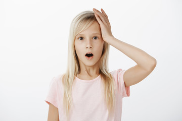 Girl forgot bag at school, remembering about it. Studio shot of forgetful cute young daughter with fair hair, holding hand on head and open mouth, recalling something in memory, feeling worried