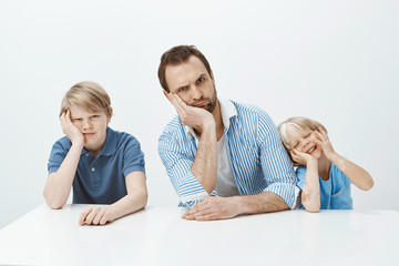 Portrait of funny family of father and sons sitting at table, leaning head on hand and making faces...