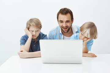 Pleased funny father sitting with sons near laptop, watching at screen with satisfied happy smile while boys feeling bored and indifferent, wanting play instead of spending time with dad