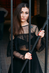 city portrait. sad girl. brunette in a black dress. expectation. dreams. portrait of a young, beautiful girl. The brunette in a black dress is standing near the cage with black rods. clings to them.