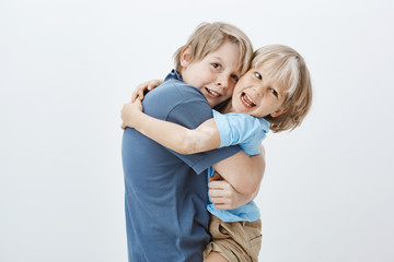 Family and positive emotions concept. Satisfied happy blond boy hugging brother and gazing at camera with pleased positive smile, being joyful while spending time with sibling over grey wall