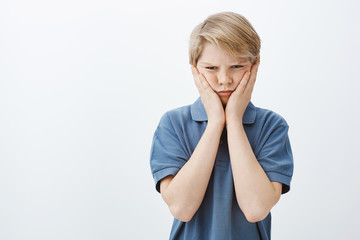 Studio shot of gloomy offended cute boy in blue t-shirt, frowning and staring unhappy at camera...