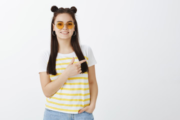 Horizontal shot of carefree friendly and stylish fashion blogger in sunglasses and trendy t-shirt, holding hand in pocket, pointing right and smiling broadly, showing direction over gray wall