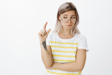 Not sure if this size fits. Unsure doubtful cute european female student with fair hair and tattoos in round glasses and trendy t-shirt, shaping something small or tiny with hand and peeking
