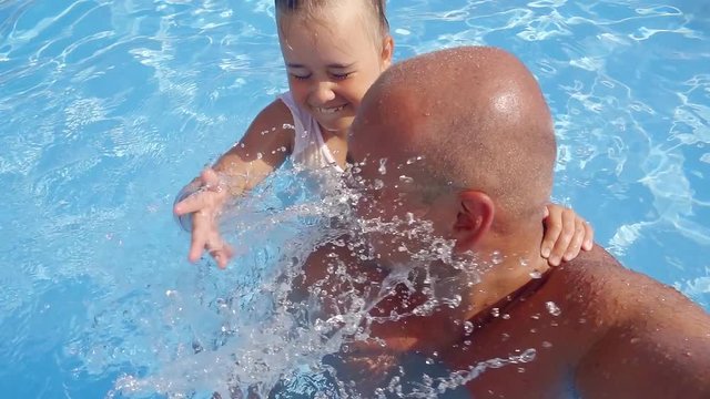 adult bald man is playing in swimming pool with his child daughter and taking video selfie, girl is splashing