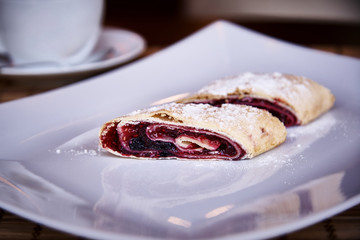 Appetizing strudel with berries on a white plate with powder and a cup on a saucer close-up.