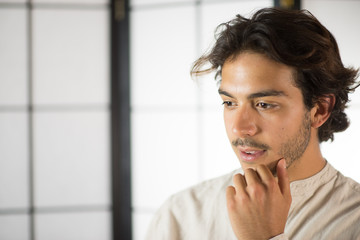 An handsome young man, 20Y, is posing in studio and touching his chin in doubt. White skirt. Retroilluminated booths in the background.