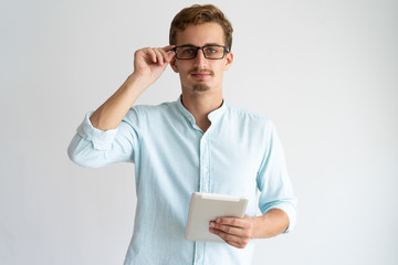 Confident intelligent man adjusting glasses and holding tablet. Serious smart young mobile app...