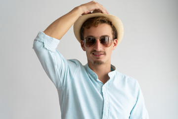 Positive excited young man in sunglasses adjusting sun hat and looking at camera. Smiling bearded guy putting on straw hat. Vacation concept