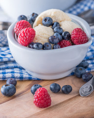 A scoop of vanilla ice cream with raspberries and blueberries on a wooden board and blue and white dish towel.
