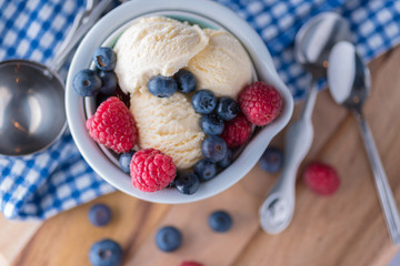 A scoop of vanilla ice cream with raspberries and blueberries on a wooden board and blue and white dish towel.
