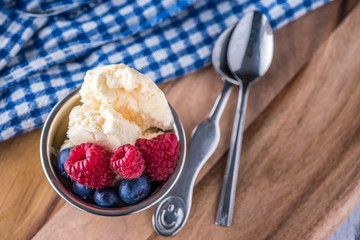 A scoop of vanilla ice cream with raspberries and blueberries on a wooden board and blue and white dish towel.
