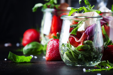 Summer salad with arugula, soft cheese, red strawberry and prosciutto in glass jars on black table, selective focus