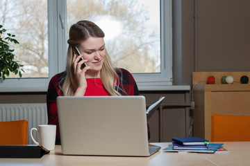 A blonde woman sitting at a laptop and talking over a cell phone.