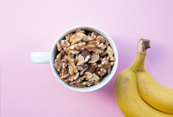 White cup full of Walnuts with banana on pink background. Above view