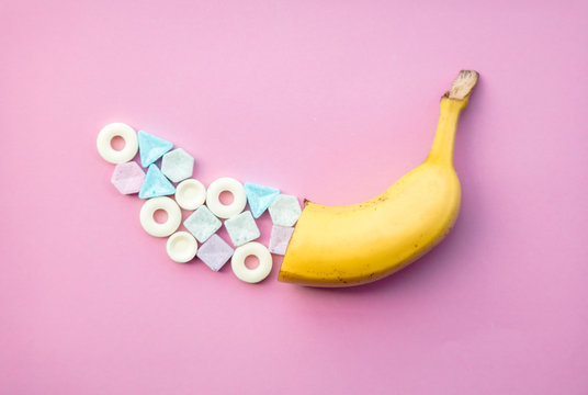 Colorful candies and half of yellow banana on pink background. Natural light, above view