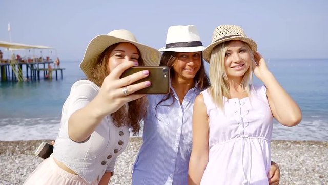 three female vacationers are taking selfie by smartphone camera, standing on a pebble beach in sunny morning