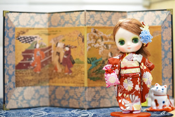 Cute doll in Kimono. A cute doll is dressing up in a red kimono. Kimono is a traditional Japanese garment. The doll is standing in Japanese setting holding omamori Japanese charm.