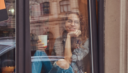 Young redhead curly girl wearing casual clothes and glasses sitting on a window sill with a takeaway coffee.