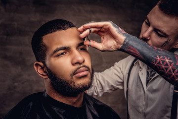 Old-fashioned professional tattooed hairdresser does a haircut to an African American client. Isolated on dark textured background.