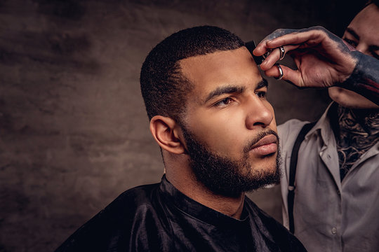 Old-fashioned professional tattooed hairdresser does a haircut to an African American client. Isolated on dark textured background.