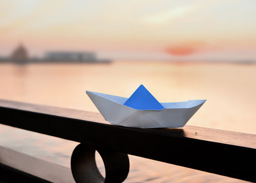 A paper boat stands on an iron black fence on the background of the river at sunset.