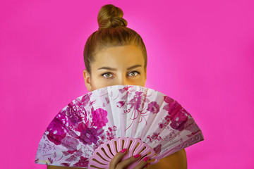 Portrait of a young beautiful girl with bright make-up and colored fan in hands close-up on the pink background. Geisha cosplay