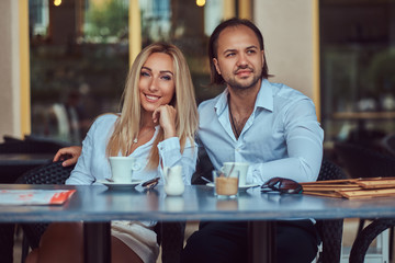 Happy attractive couple - charming blonde female dressed in a white blouse and bearded male with a stylish haircut dressed in a white shirt during a date at a cafe outdoors.