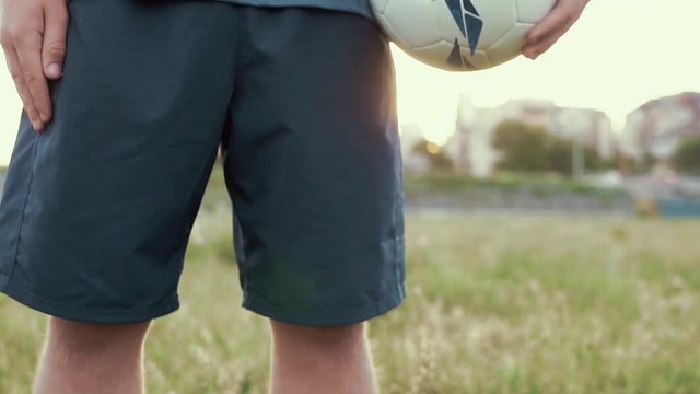 Slide slow motion a football player's figure with a ball in his hand on his side. A young athlete stands on a football field before the match at sunset on a sunny day in the summer. The lights of sun