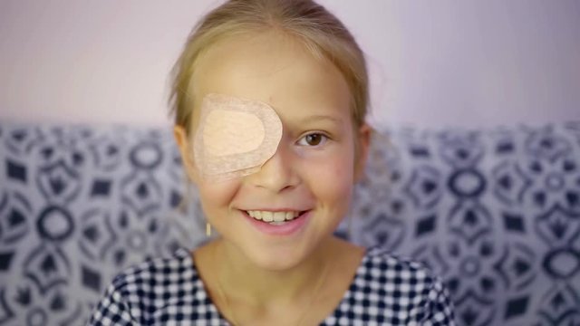 cheerful little girl is sitting in a room and smiling for camera, having protective bandage on one eye