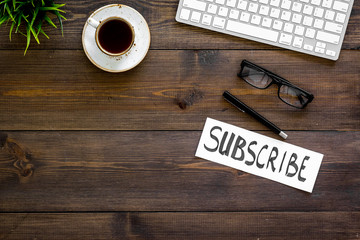 Email subscribe concept. Hand lettering subcribe on work desk with plant, glasses, computer keyboard, cup of coffee on dark wooden background top view copy space
