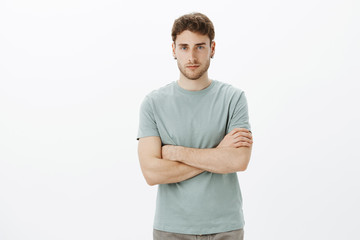 Indoor shot of confident good-looking european guy with fair hair in casual t-shirt, holding hands crossed on chest and looking with serious expression at camera, being self-assured and determined