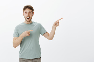 Pleased surprised attractive guy with fair hair, dropping jaw while discussing something amazing, pointing right with index fingers, gasping from joy and excitement, standing over gray background