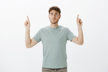 Worried intense funny fair-haired guy in t-shirt, pointing up with raised index fingers and looking at sky with anxious scared expression, worrying for child on playground, standing over gray wall