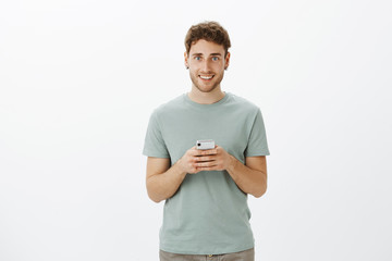 Cute friendly european fair-haired guy with bristle in casual clothes, holding smartphone, smiling broadly at camera, hoping to find love in new app for his brand new device, standing over grey wall