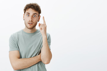 Waist-up shot of energetic good-looking european guy in earrings and t-shirt, raising index finger in eureka gesture and staring at camera, finally having great idea or plan, standing over grey wall - obrazy, fototapety, plakaty