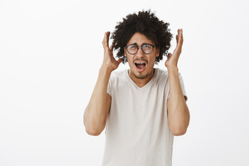 Portrait of pissed and irritated handsome young male student in glasses, scraeming from pressure and stress, shaking hands near head, frowning, being annoyed and fed up of everything over gray wall