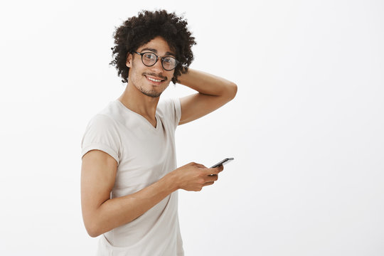 Good-looking friendly male gay with bristle and moustache, scratching head and smiling broadly, turning at camera while standing in profile, using smartphone, posing over gray background
